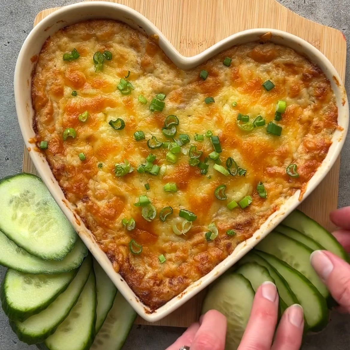 A heart-shaped Easy Tuna Melt Dip with Cheddar casserole topped with melted cheese and chopped green onions, surrounded by sliced cucumbers on a wooden board.