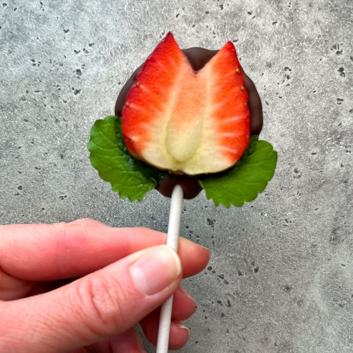 A hand holds a lollipop shaped like a tulip, made from a halved strawberry dipped in chocolate with two green leaves at the base—a creative bite perfect for health-conscious food recipes, against a gray textured background.
