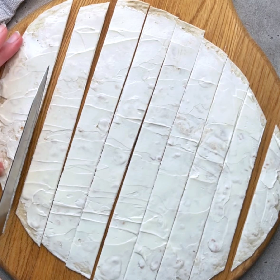 A tortilla topped with cream cheese is being cut into strips with a knife.