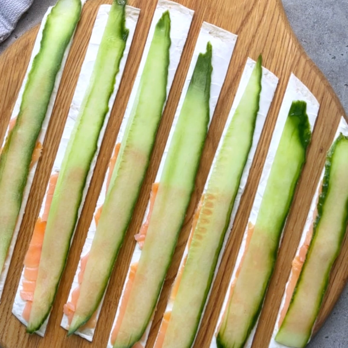 Seven tortilla strips are topped with smoked salmon and cucumber and arranged on a wooden chopping board.