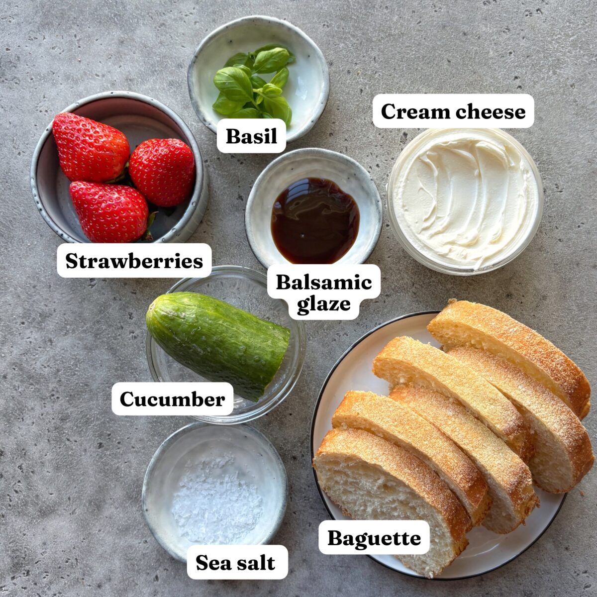 Sliced baguette, cream cheese, strawberries, cucumber, basil, balsamic glaze, and sea salt are arranged in separate bowls on a gray surface.