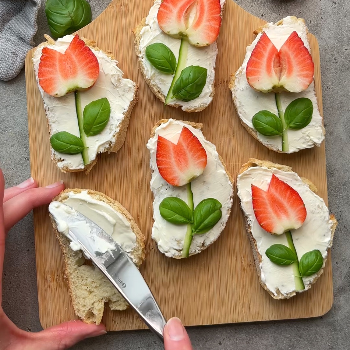 Six slices of bread with cream cheese and strawberry tulip decorations garnished with basil leaves, on a wooden board. A hand spreads cream cheese on a slice in the foreground.