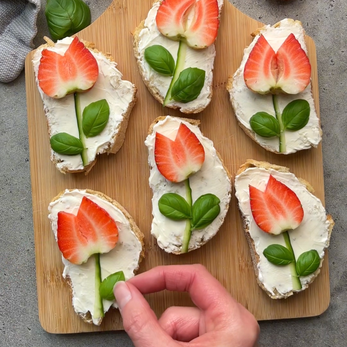Six slices of bread with cream cheese are arranged on a wooden board, each transformed into strawberry crostini by topping them with sliced strawberries and basil leaves to resemble flowers. A hand is placing a basil leaf on one slice.