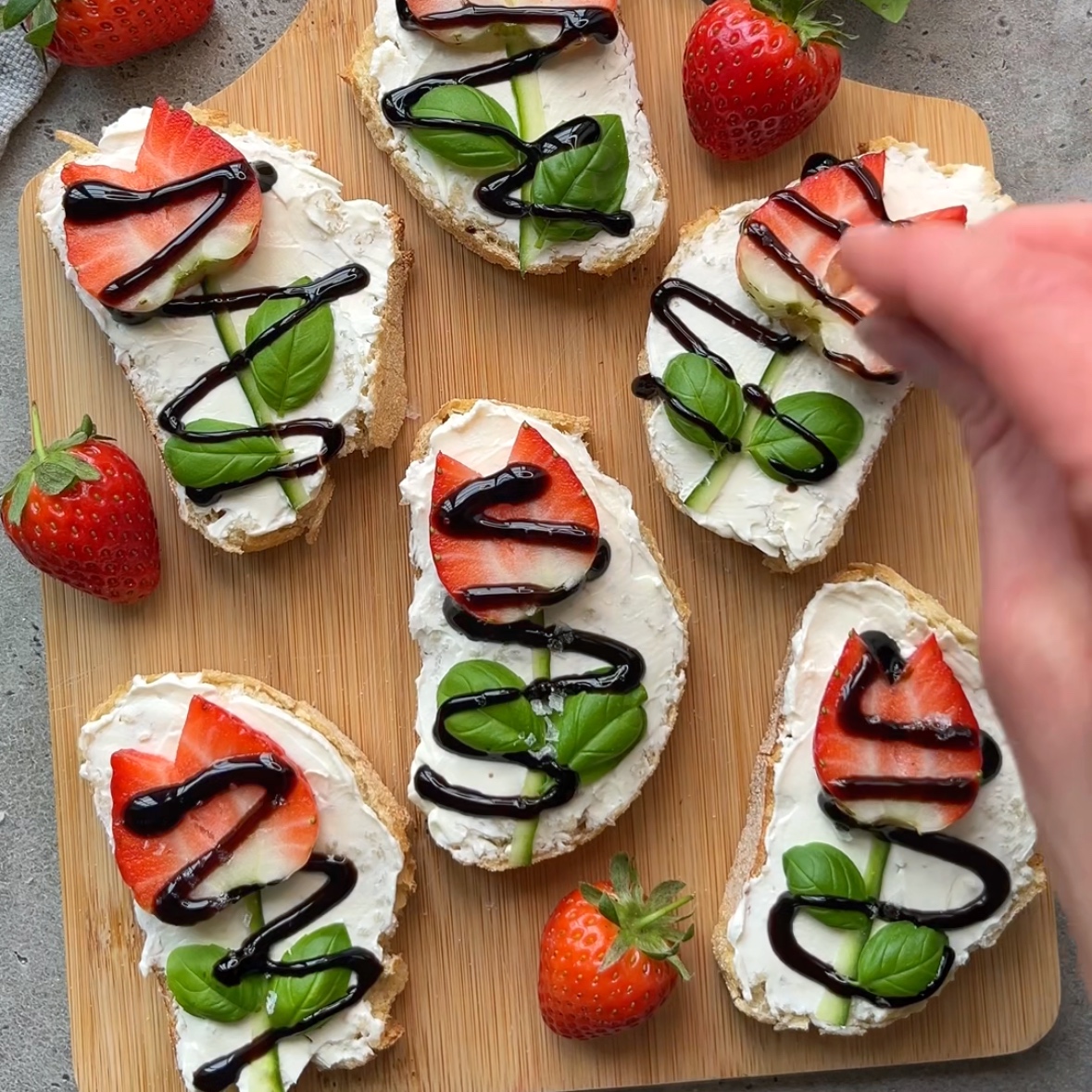 Slices of bread with cream cheese, strawberries, cucumber, basil leaves, and balsamic glaze on a wooden board. A hand adds glaze to one slice. Whole strawberries are scattered around.