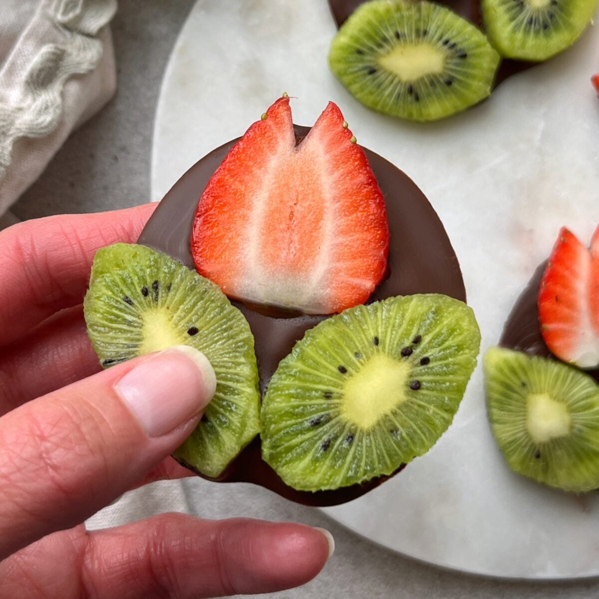 A hand holding a chocolate-covered disc topped with a slice of strawberry shaped into a flower and two slices of kiwi shaped into leaves.