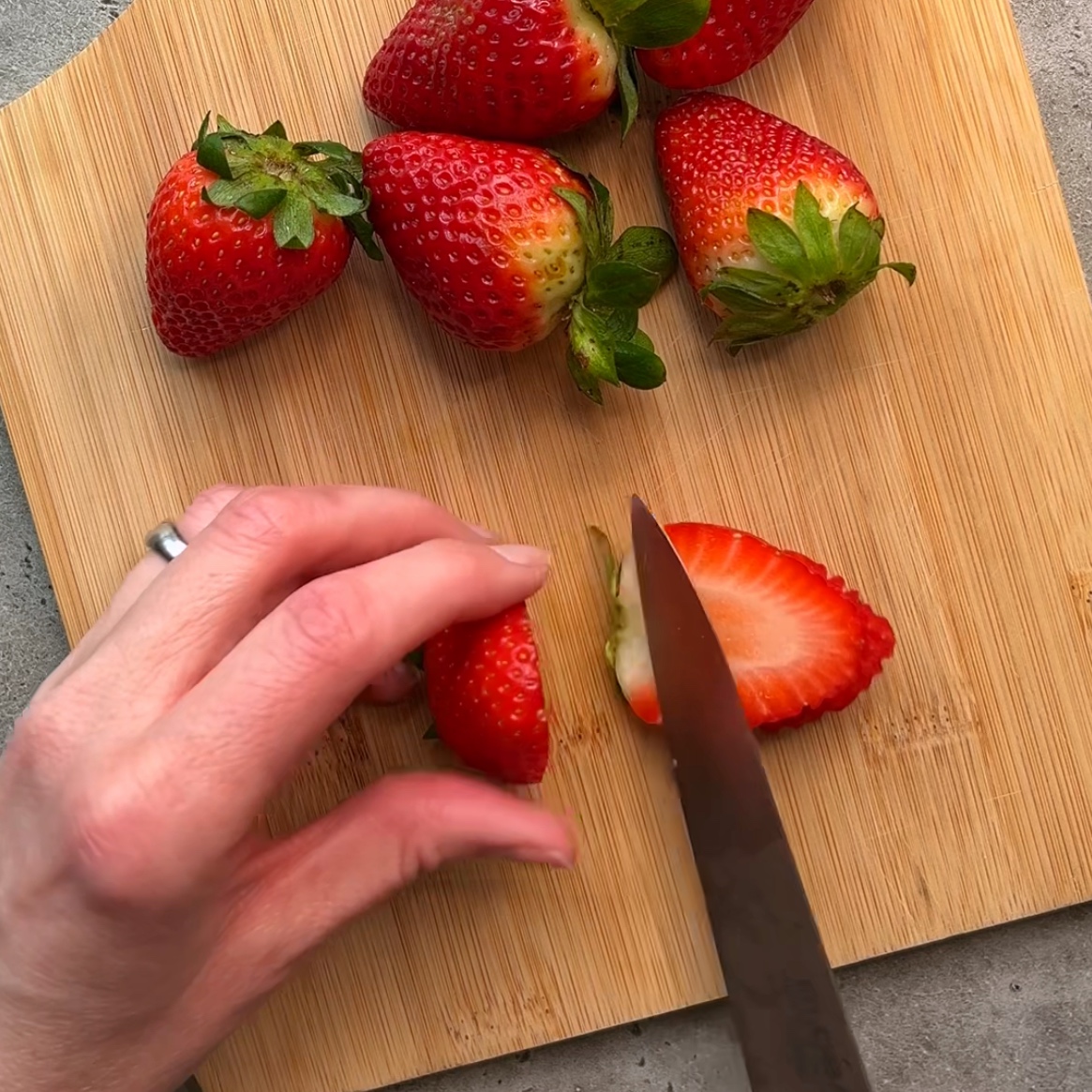 A hand slices a strawberry with a knife on a wooden cutting board.