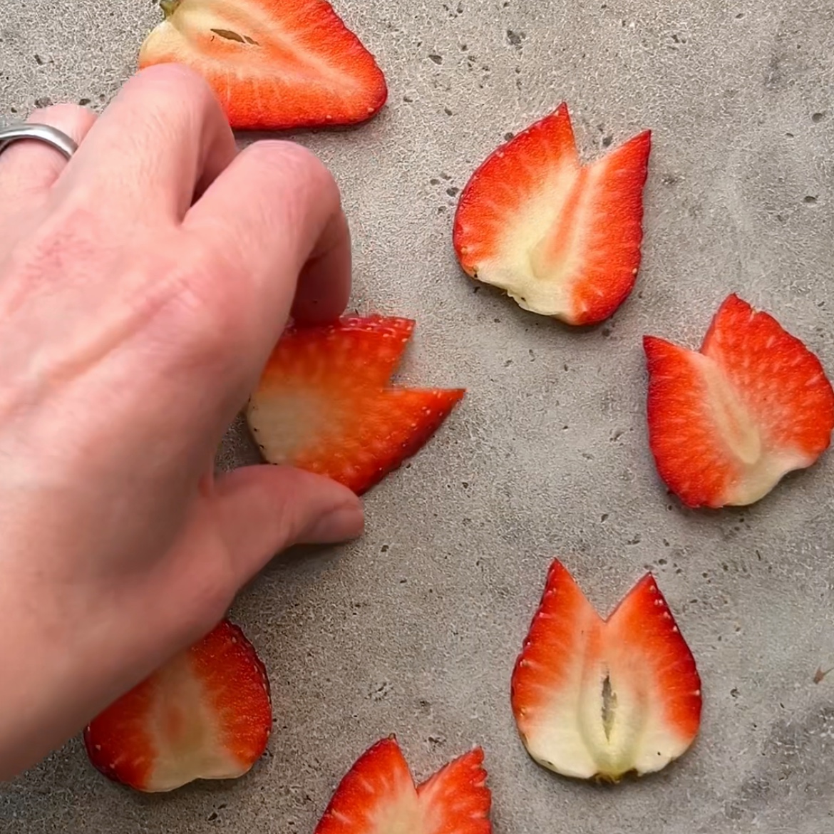 A hand arranges strawberry slices shaped like tulip flowers on a gray surface.