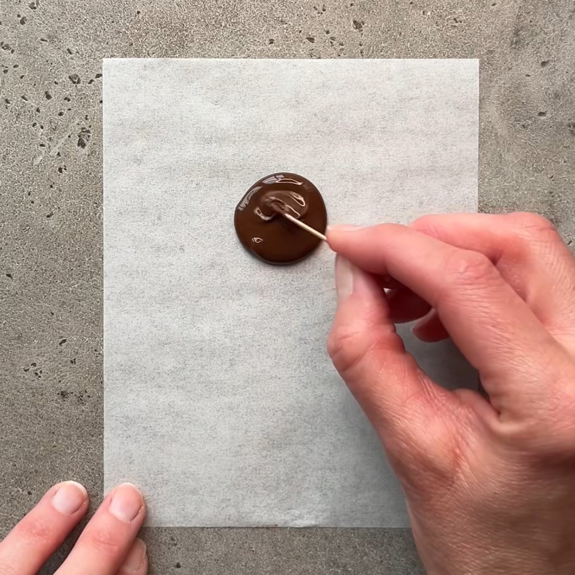 A hand uses a toothpick to swirl melted chocolate on parchment paper.