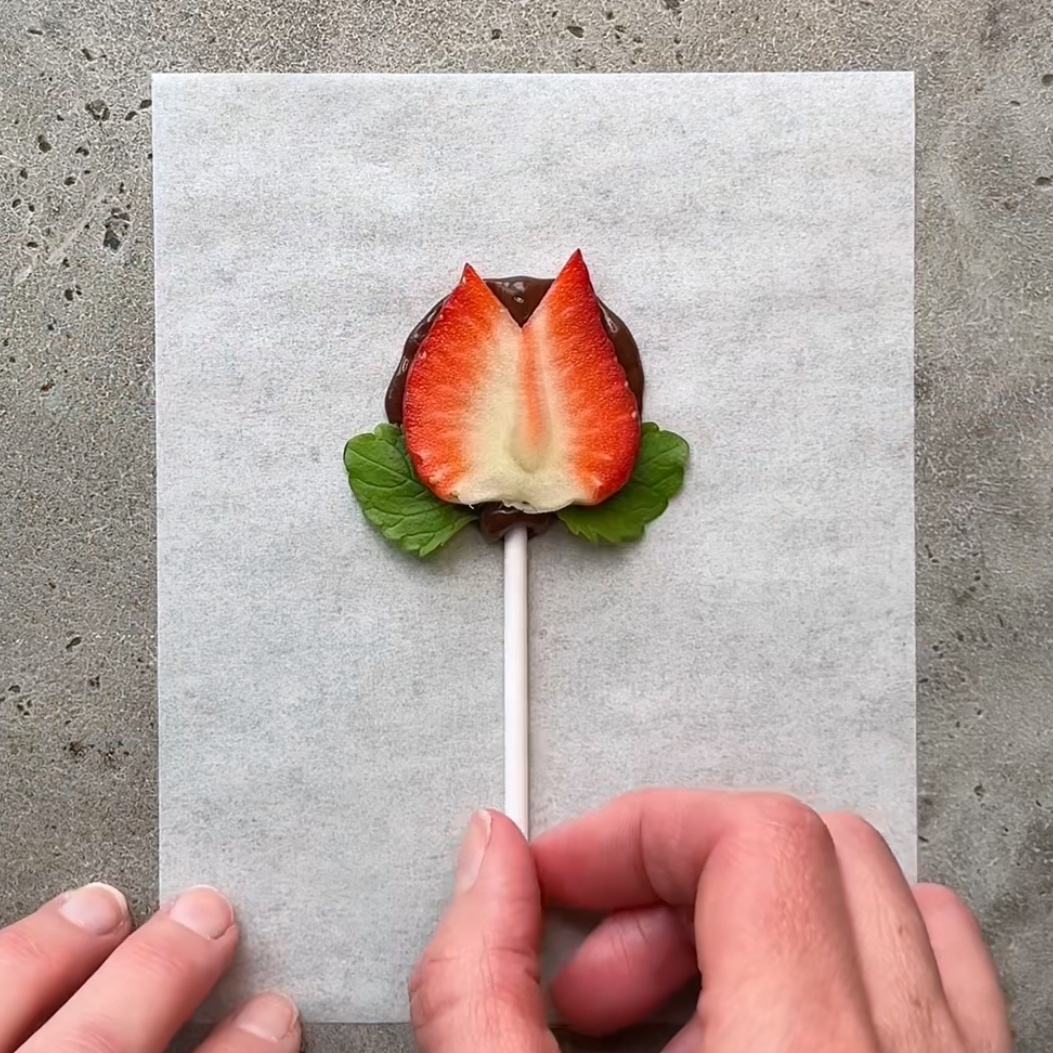 A chocolate-covered treat on a stick with a strawberry slice shaped like a tulip and small green leaves.