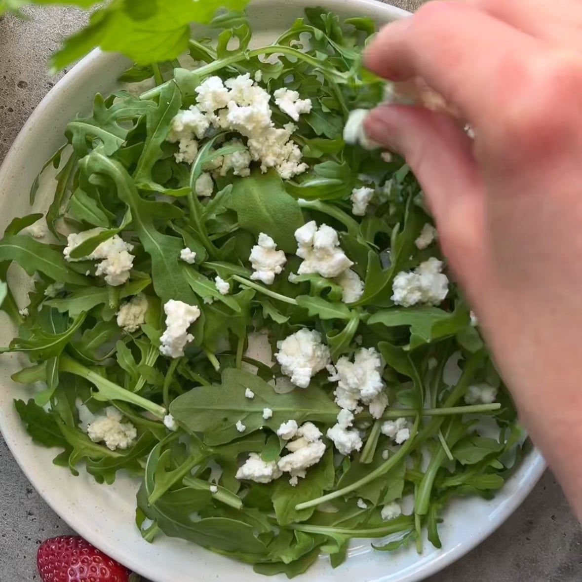 A hand sprinkles crumbled cheese over a plate of fresh arugula leaves.