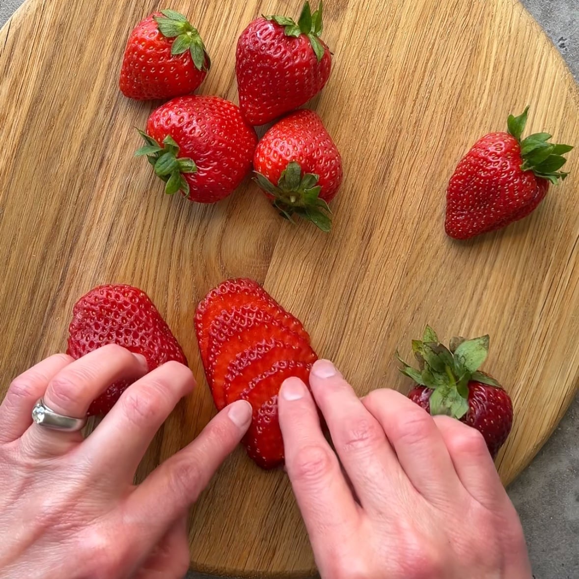 Hands slicing a strawberry on a wooden cutting board, with whole strawberries scattered around.