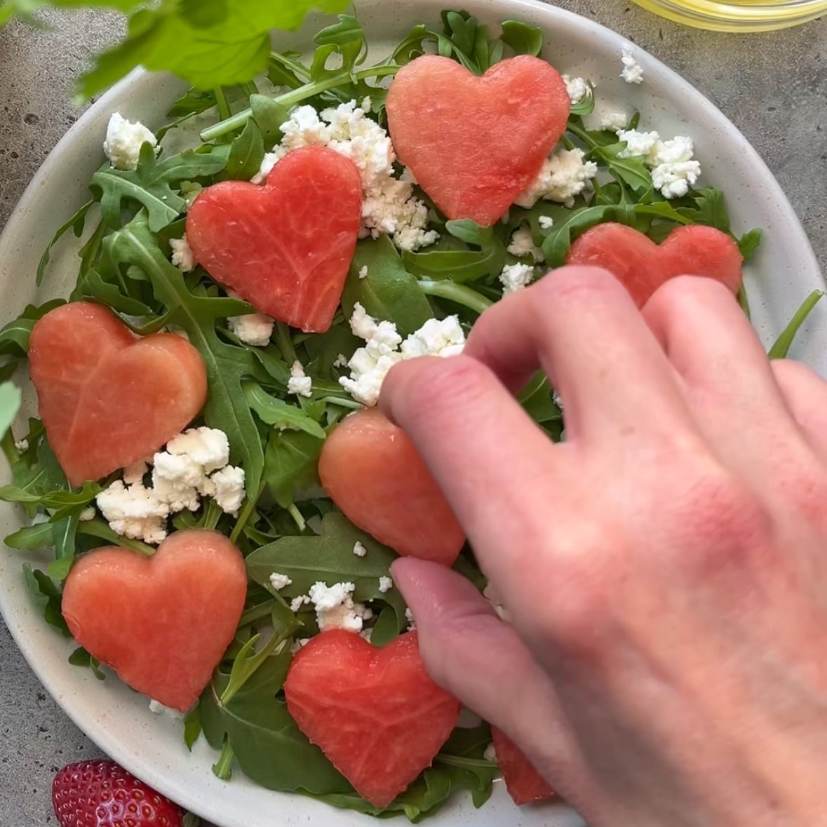A hand places heart-shaped watermelon slices on a bed of arugula and crumbled cheese in a white plate.