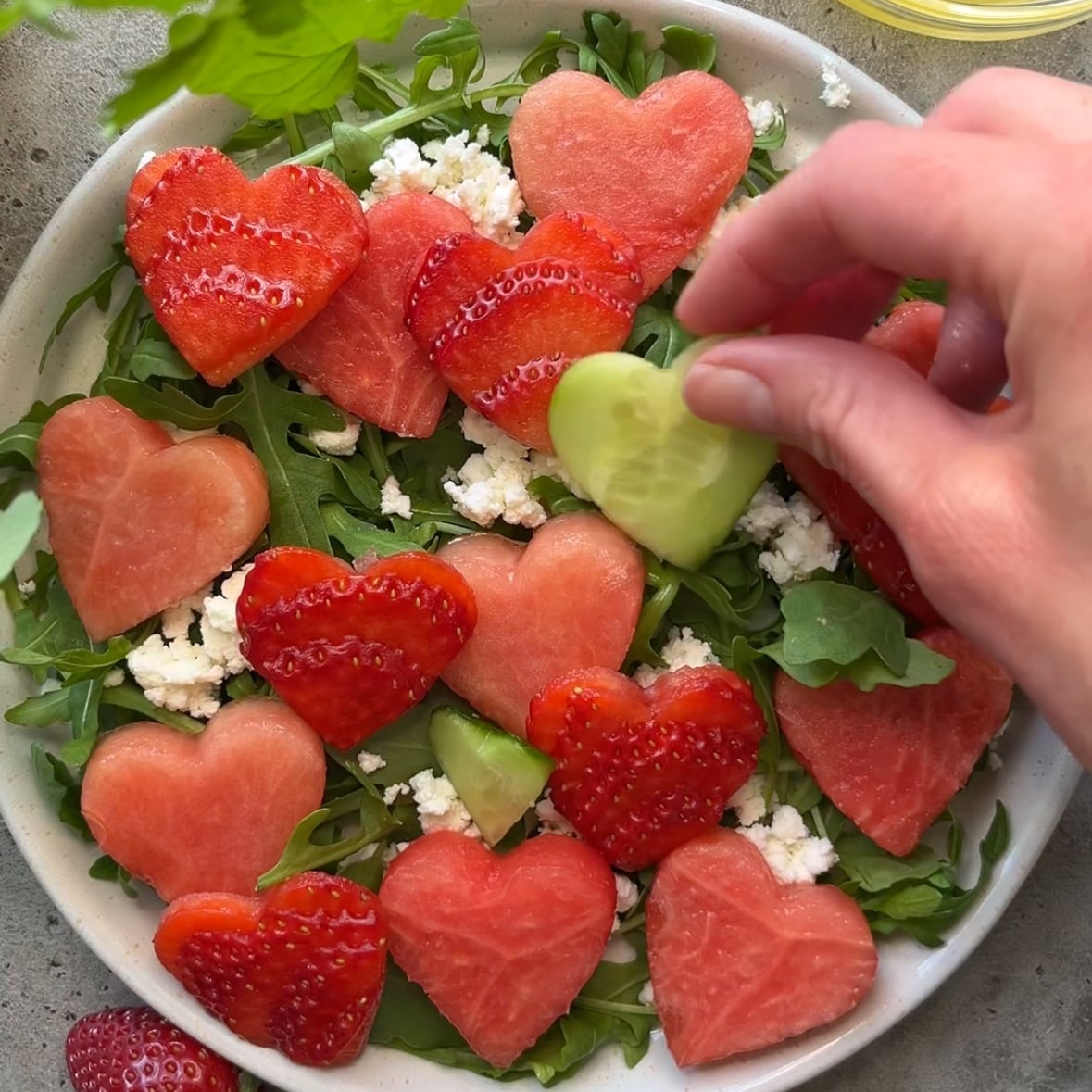 A hand places a heart-shaped cucumber slice on a beautifully arranged Strawberry Watermelon Salad, featuring heart-shaped watermelon, strawberries, arugula, and crumbled cheese.