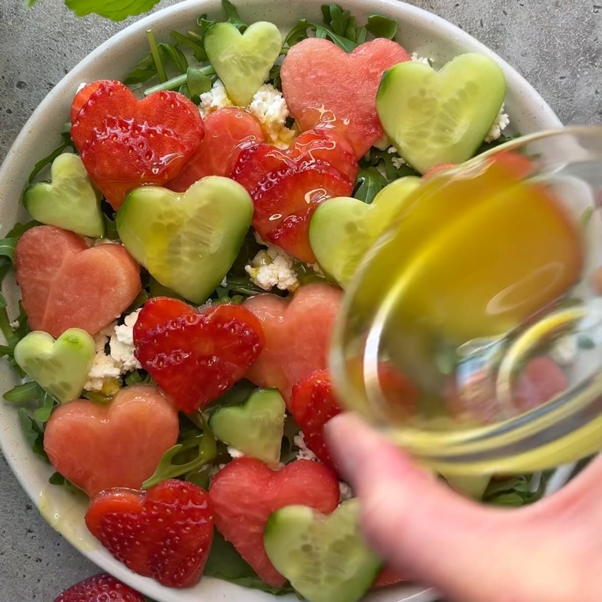 A Strawberry Watermelon Salad features heart-shaped slices of cucumber, watermelon, and strawberry, all topped with crumbled cheese as an olive oil dressing is poured over it.