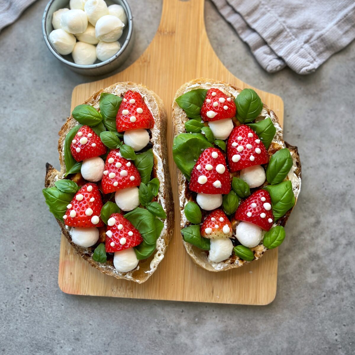 Two slices of bread topped with fresh basil leaves, mozzarella balls, and halved strawberry toadstools, arranged on a wooden board with more mini mozzarella in a bowl nearby.