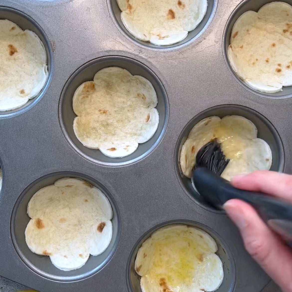 A hand brushes olive oil onto flower-shaped tortillas placed in a muffin tin.