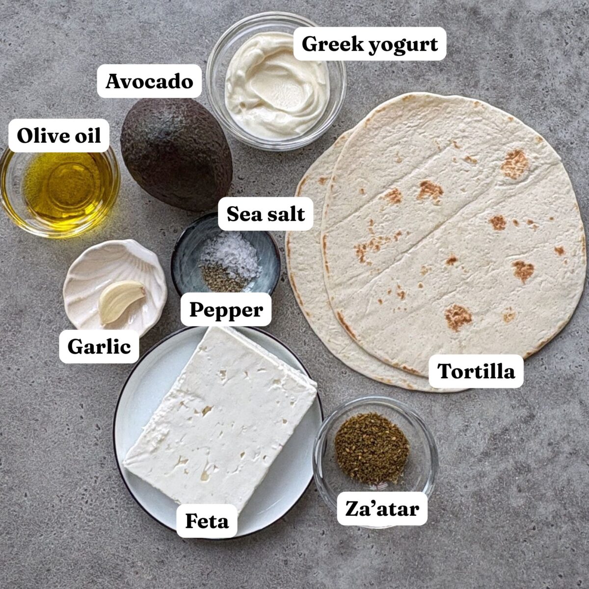 Ingredients on a gray surface: avocado, Greek yogurt, olive oil, garlic, sea salt, pepper, feta cheese, zaatar, and tortillas.