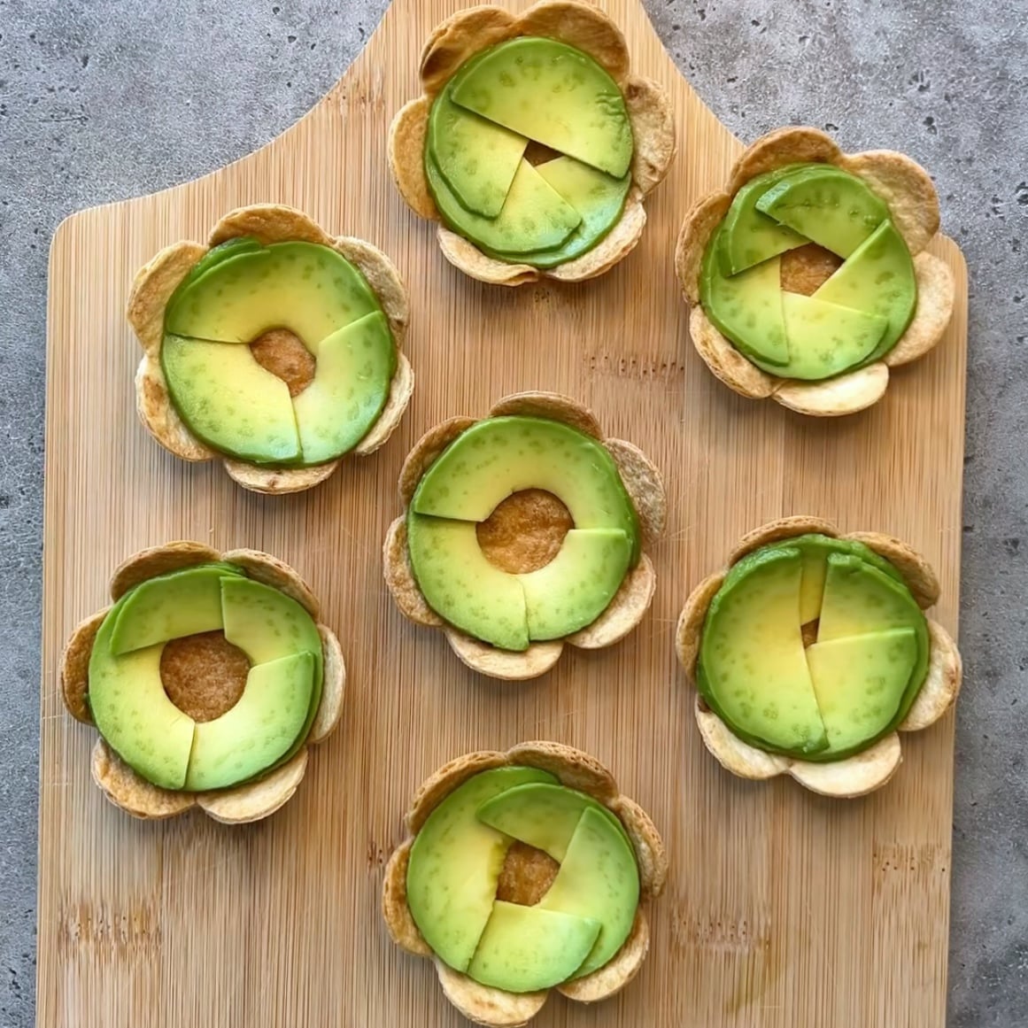 Seven avocado and flower-shaped tortilla appetizers are arranged on a wooden cutting board.