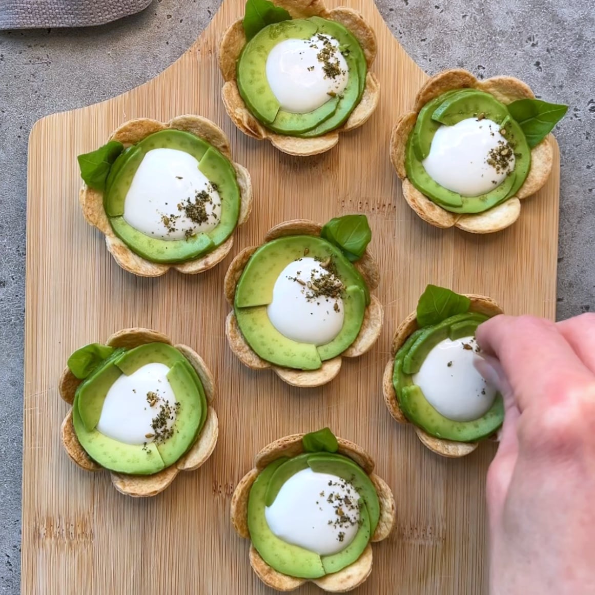 Seven flower-shaped tortilla appetizers with sliced avocado, whipped feta, herbs, and basil leaves on a wooden board. A hand is placing a leaf on one appetizer.