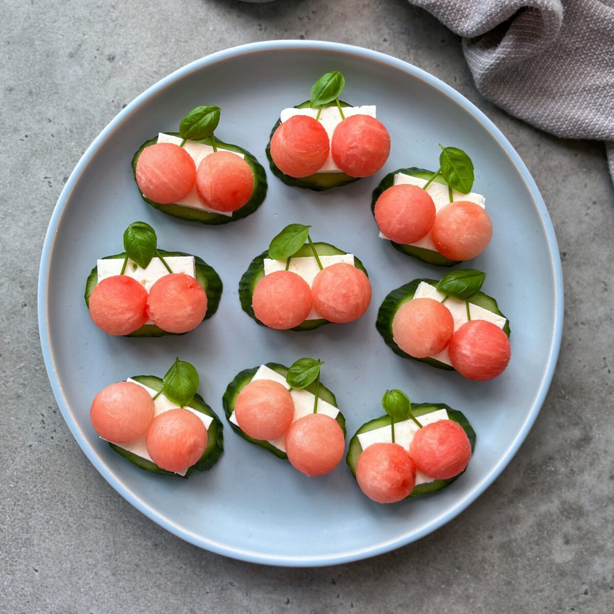 A round plate with nine bites made of two watermelon balls, a slice of feta, and a basil leaf on a cucumber slice, arranged on a gray surface.