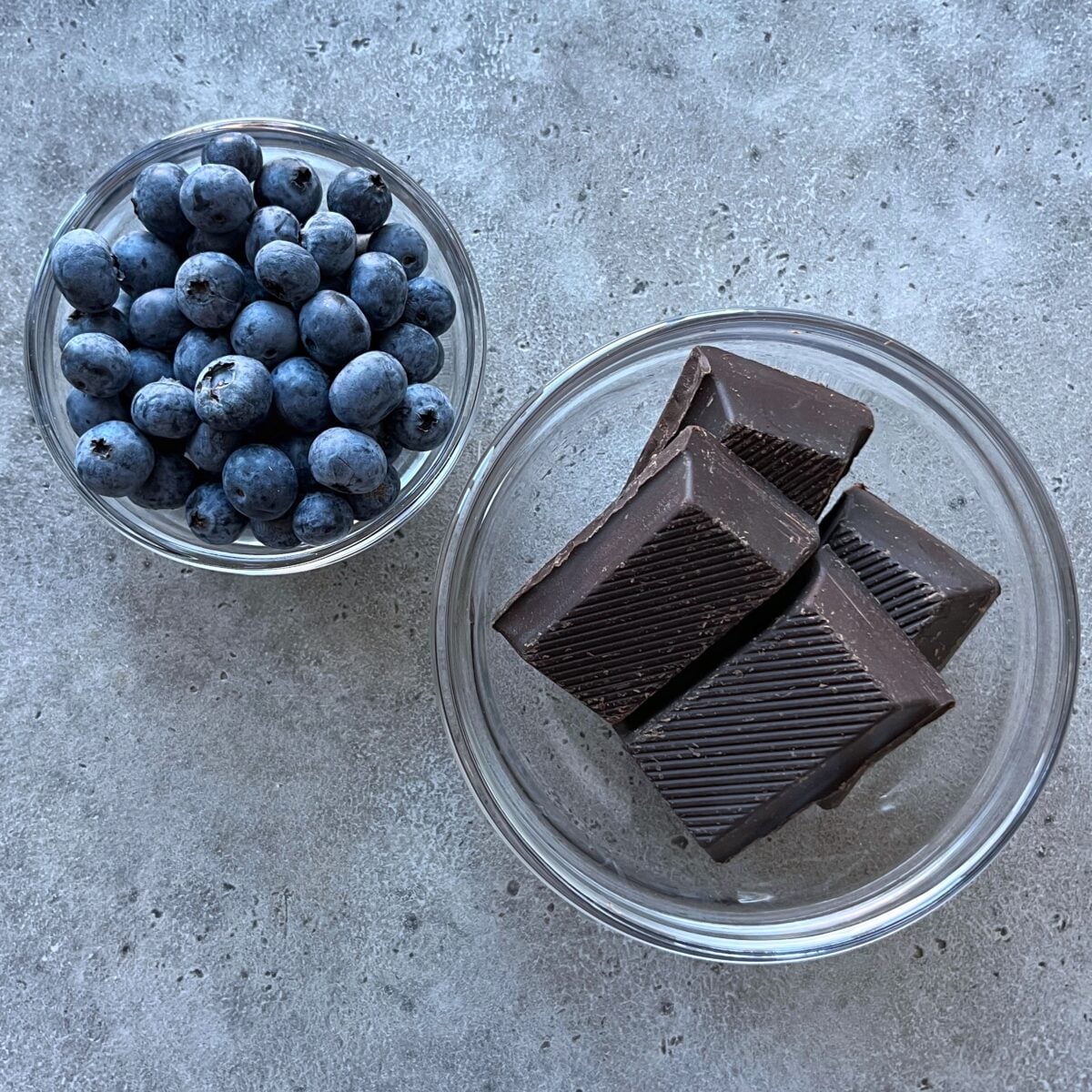 Two glass bowls on a gray surface, one brimming with blueberries and the other holding four pieces of dark chocolate.