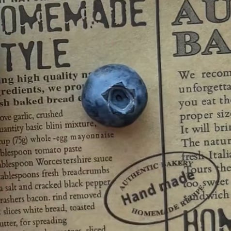 A single blueberry rests on a vintage-style parchment paper with printed text and a handmade bakery stamp.