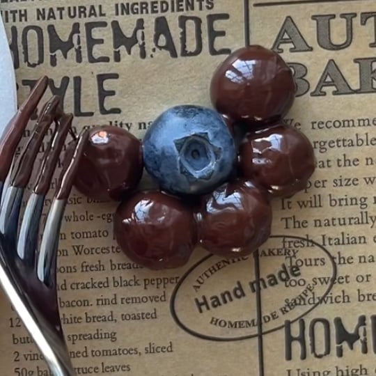 A single blueberry, surrounded by five chocolate covered blueberries, is arranged on brown parchment wrapping paper with a fork pushing one of the berries into place. 