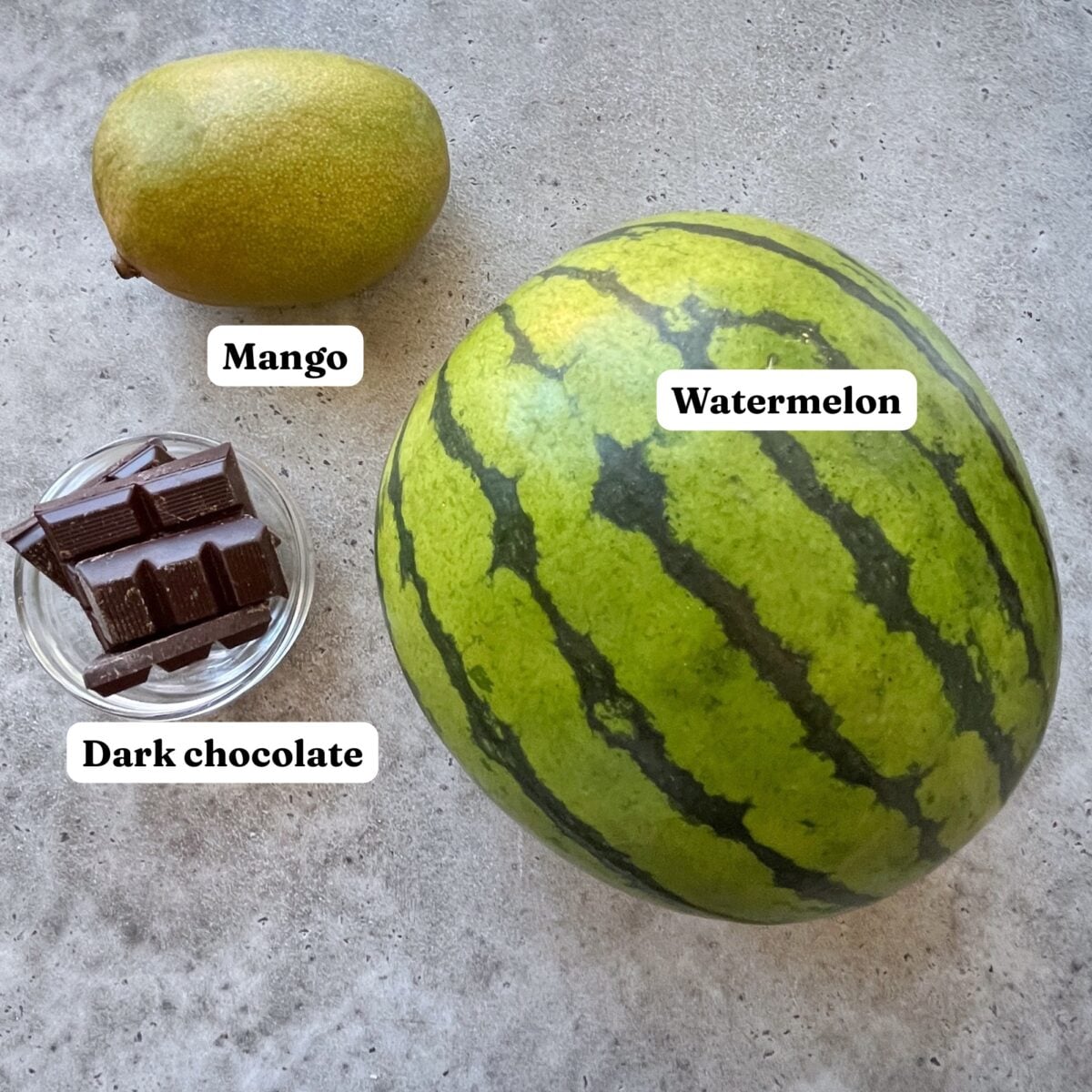 A whole watermelon, a mango, and a small bowl of dark chocolate pieces are labeled on a gray surface.