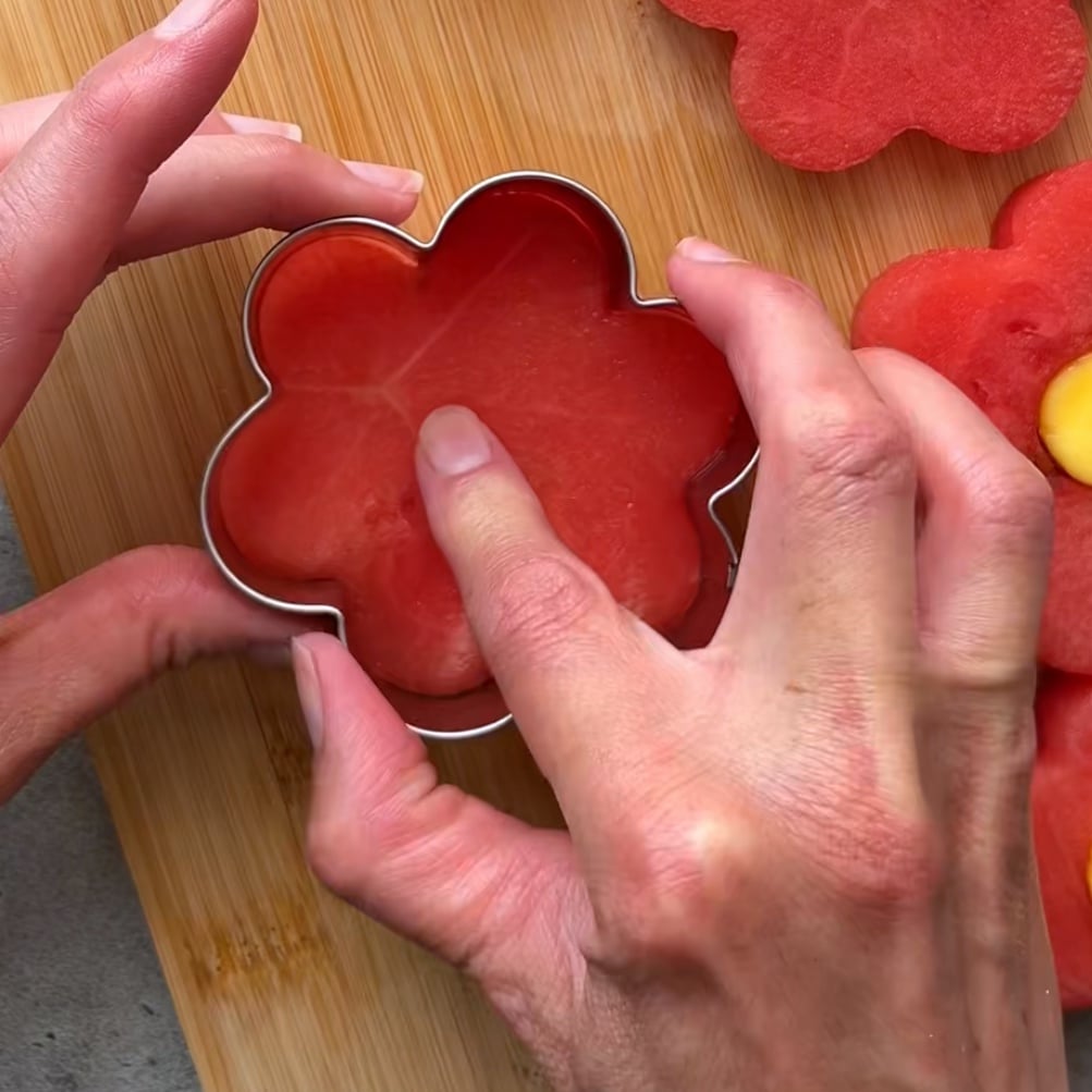 A person presses a flower-shaped cookie cutter into a slice of watermelon on a wooden cutting board.