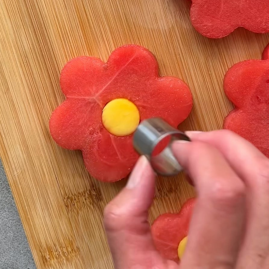 A hand places a small mango disc in the center of a flower-shaped watermelon slice on a wooden cutting board.
