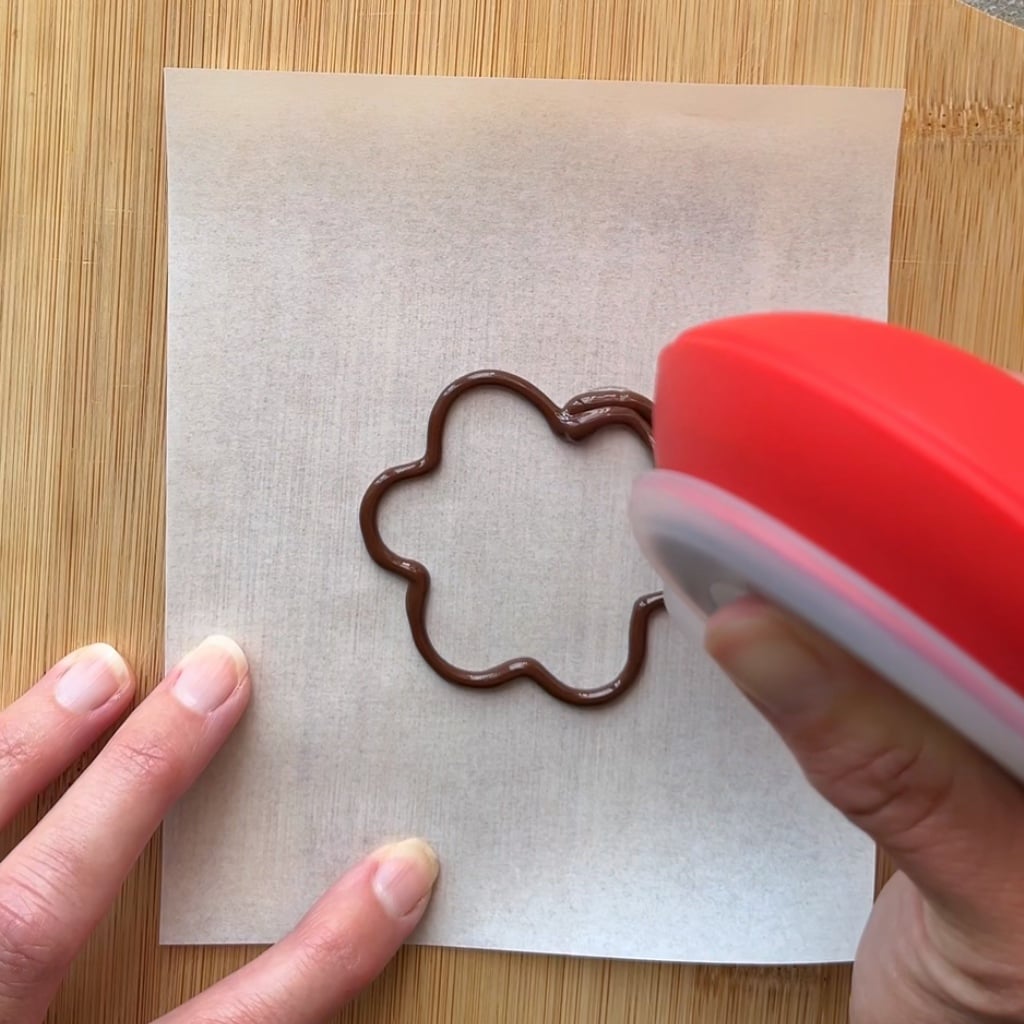 A hand holds a red device piping chocolate in a flower shape onto parchment paper on a wooden surface.