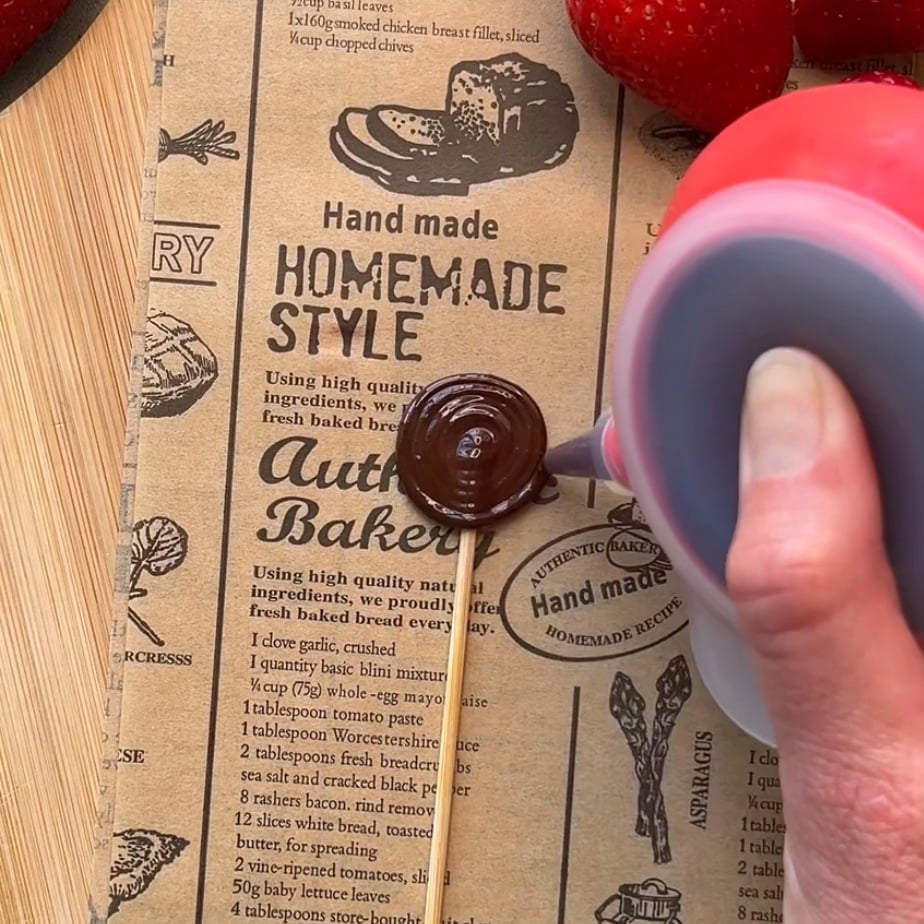 A hand pours chocolate from a squeeze bottle onto printed parchment paper with bakery-themed designs and text.