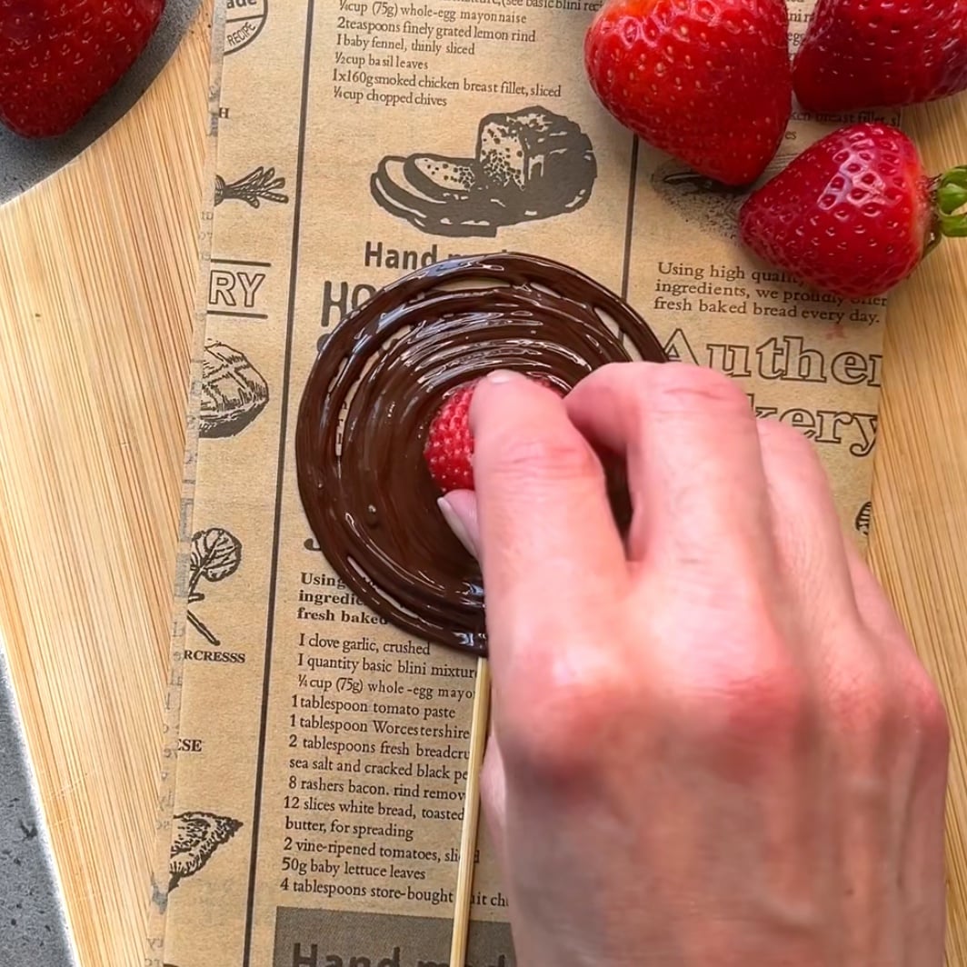 A hand dips the tip of a strawberry into melted chocolate on a skewer.