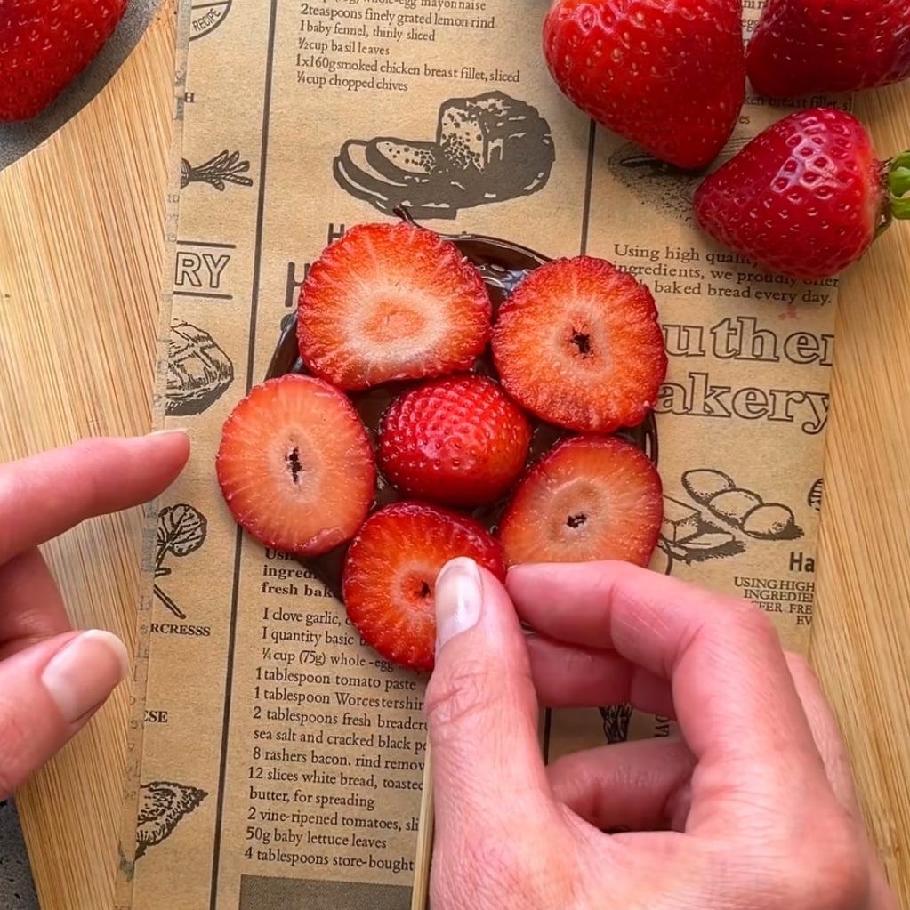 Hands placing sliced strawberries in a circular pattern on newspaper-print parchment.