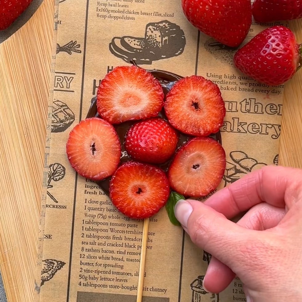 A hand arranges a basil leaf on a chocolate strawberry design, over a vintage-style newspaper on a wooden surface.