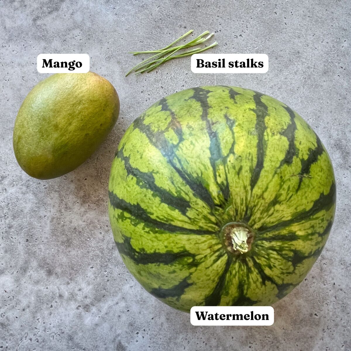 A mango, basil stalks, and a watermelon are arranged on a gray surface.
