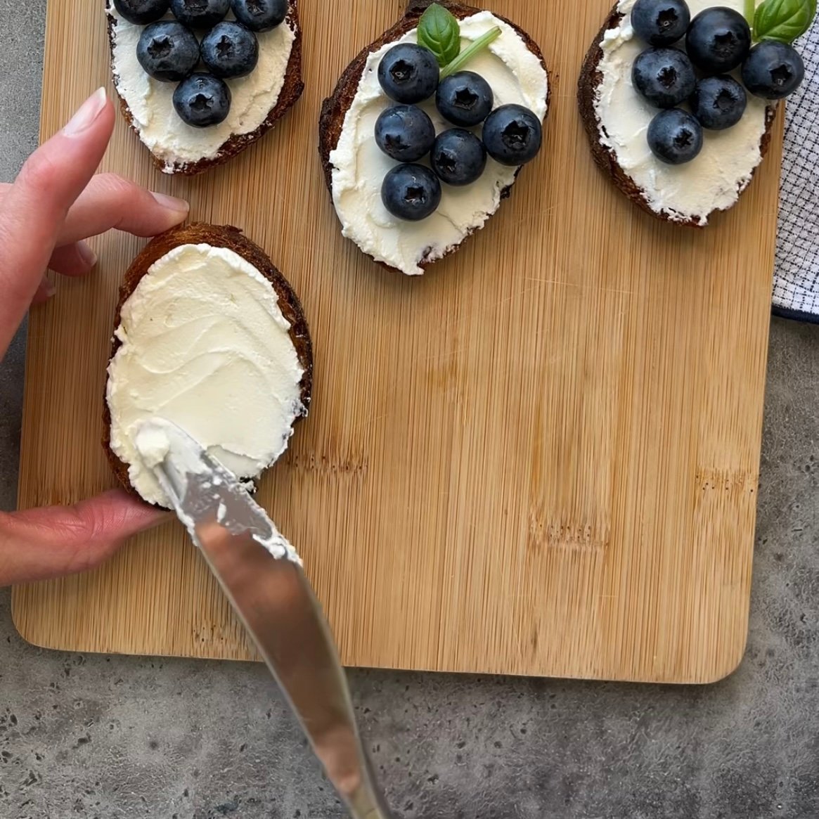 A hand spreads cream cheese on a slice of bread next to three slices topped with goat cheese with honey appetizer.