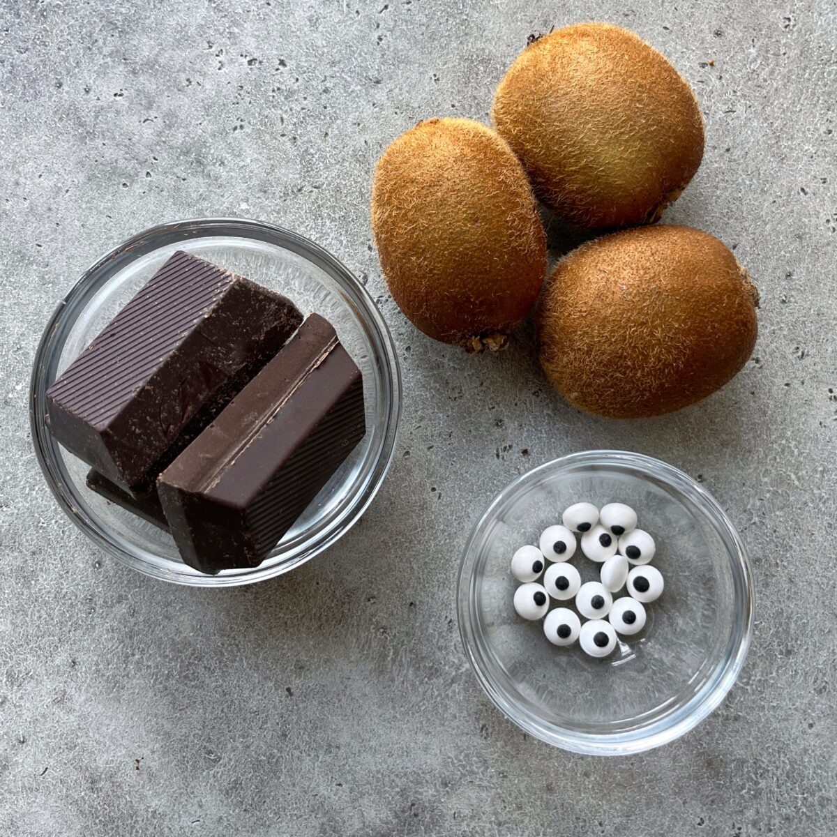 Three whole kiwis, two pieces of dark chocolate, and a small bowl of white candy eyeballs are arranged on a gray surface.