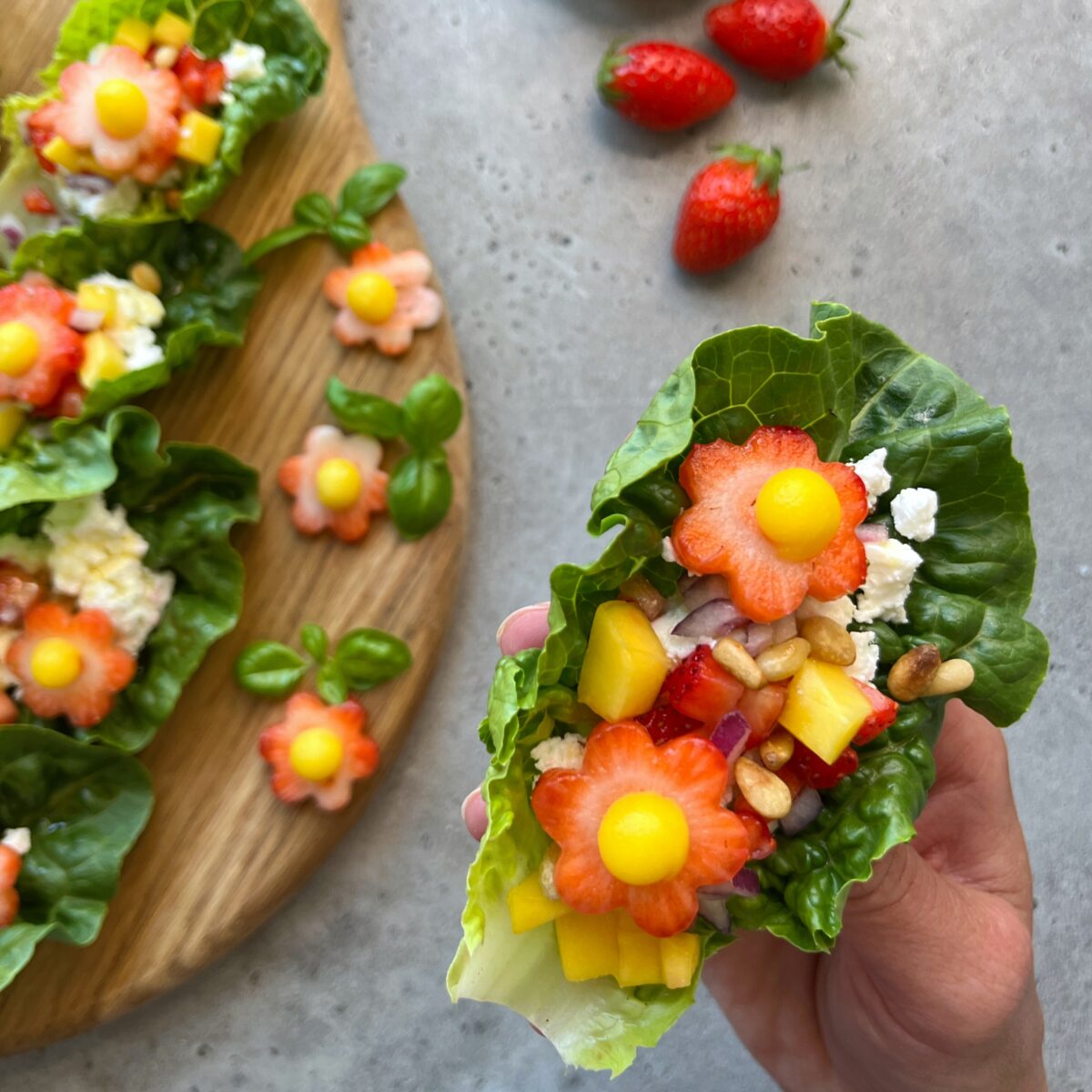 A hand holds a lettuce wrap with feta topped with flower-shaped strawberry and mango. More wraps are on a wooden board nearby.