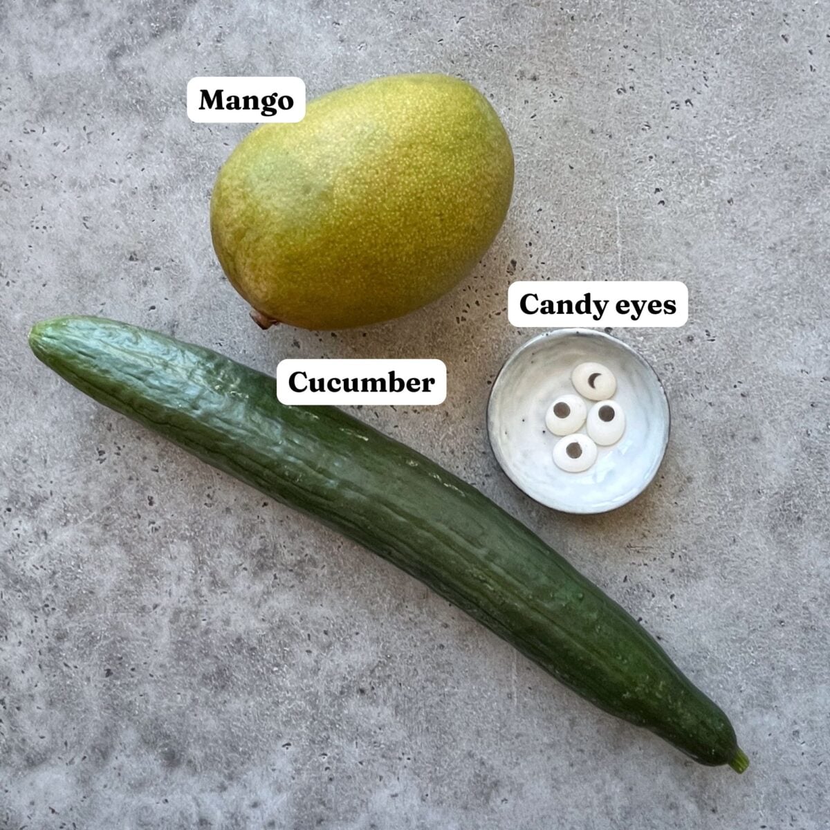 A mango, a cucumber, and a small dish of candy eyes are arranged on a gray, textured surface.