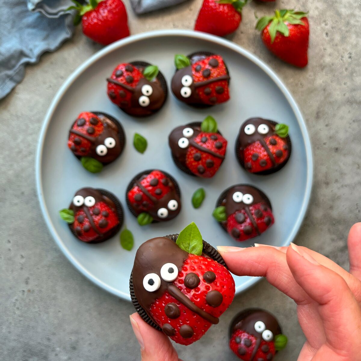 A hand holds a chocolate-covered cookie decorated as a ladybug with strawberries, candy eyes, and chocolate dots. In the background, a plate offers more tasty bites and fresh strawberries.