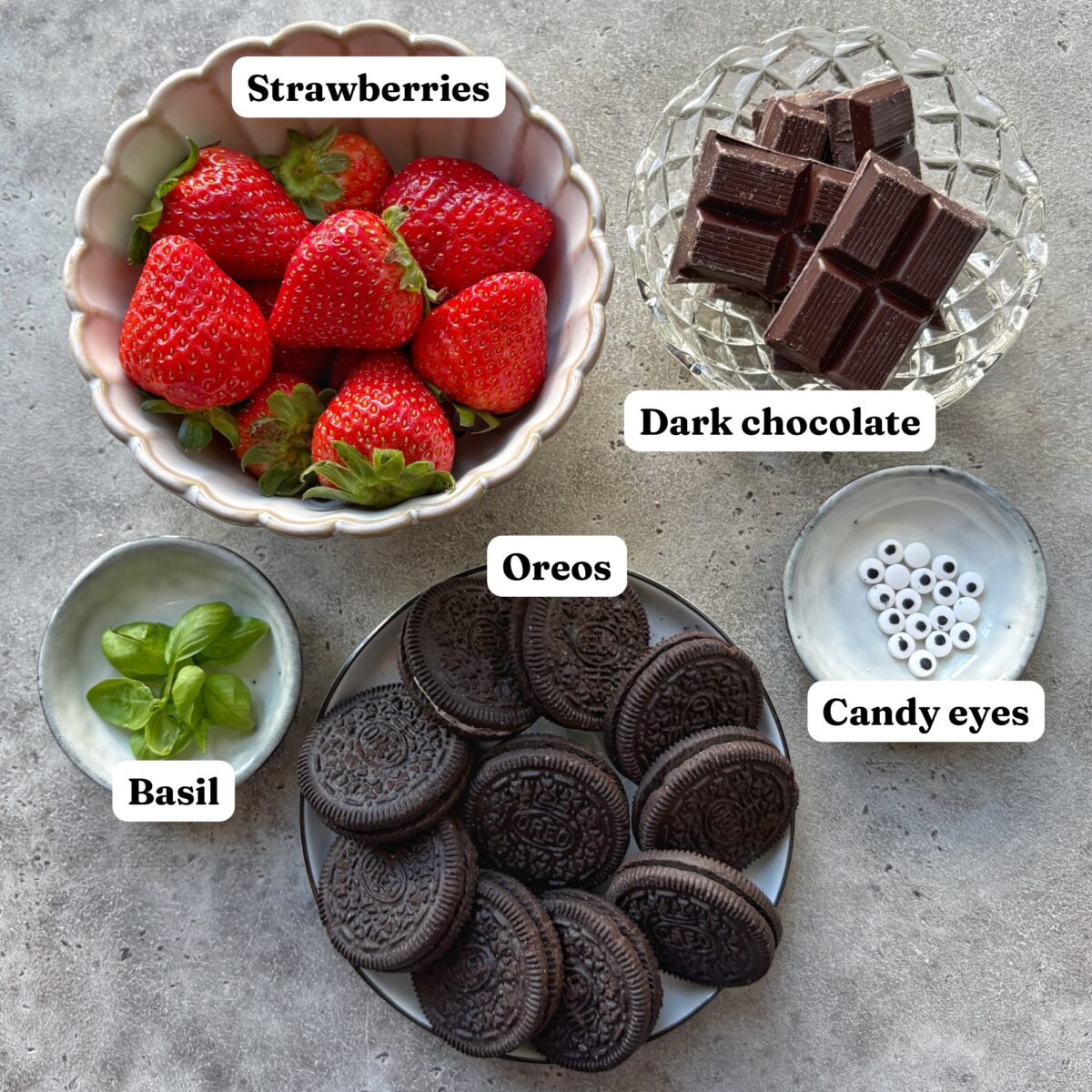 Five bowls contain labeled ingredients for making Strawberry Ladybugs with Oreos including fresh strawberries, pieces of dark chocolate, basil leaves, Oreo cookies, and candy eyes, all arranged on a gray surface.
