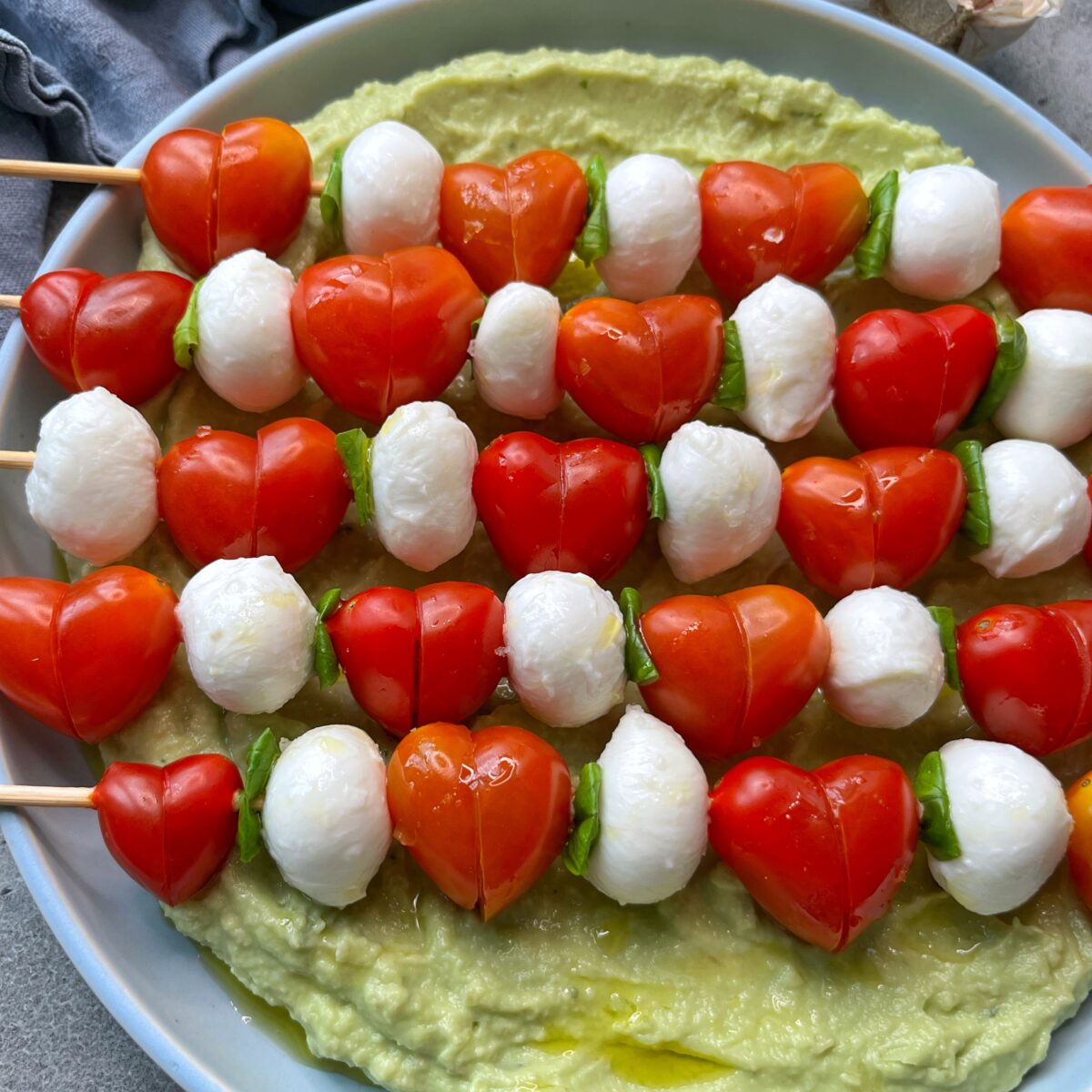 Skewers with heart-shaped grape tomatoes, mozzarella balls, and basil leaves arranged on a plate of avocado cream.