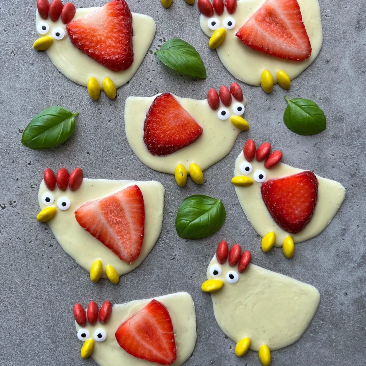 White chocolate chickens decorated with strawberries, mini yellow M&Ms as feet, and candy eyes arranged on a gray surface.