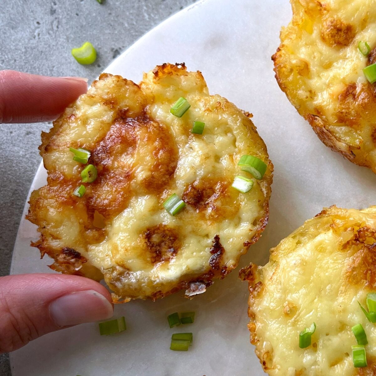 A hand holding a baked cheese smashed potato  garnished with chopped green onions.