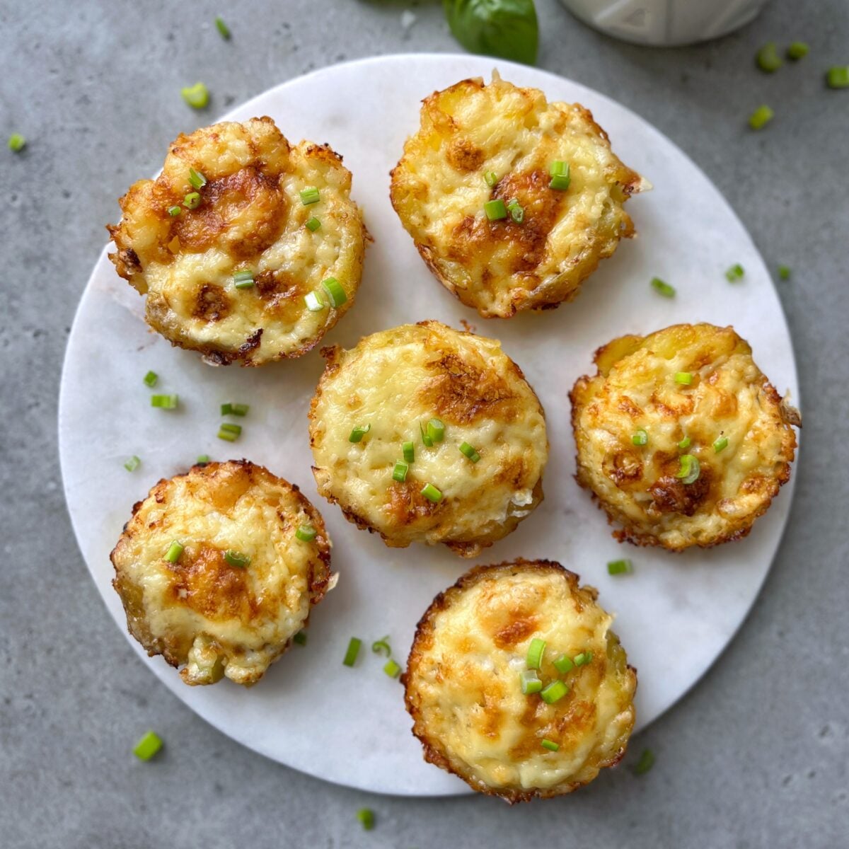 Six smashed potatoes topped with melted cheese and chopped chives are arranged on a round white platter.