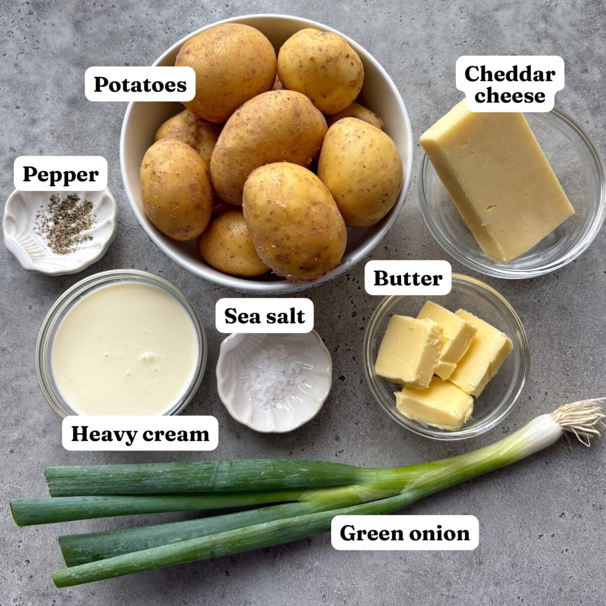 Top view of cheesy smashed potatoes ingredients featuring potatoes, cheddar cheese, butter, heavy cream, green onion, sea salt, and pepper arranged in separate labeled containers on a gray surface.