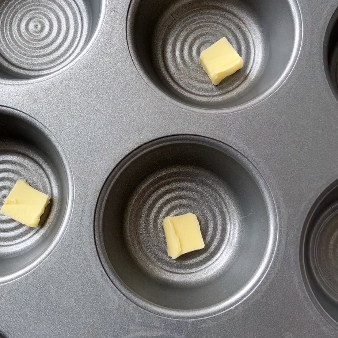 A muffin tin with several cups, three of which contain small cubes of butter in the center.