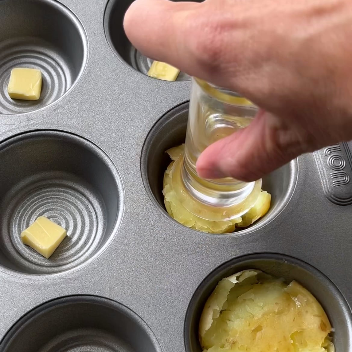 A hand uses a glass to press down a boiled potato in a muffin tin.