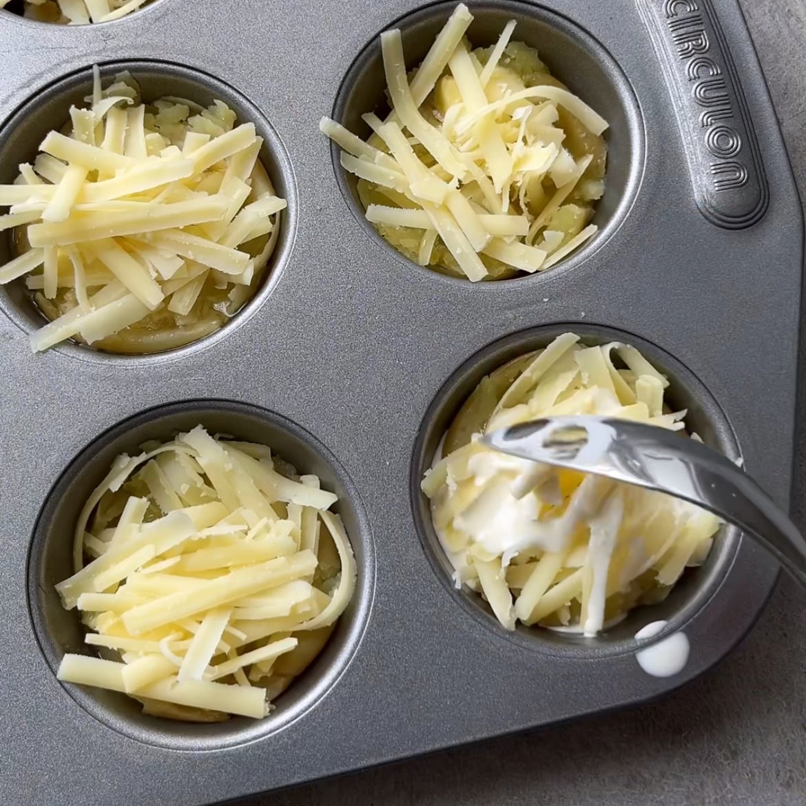 A muffin tin filled with cheesy smashed potatoes and cheese, with a spoon adding cream to one of the compartments.